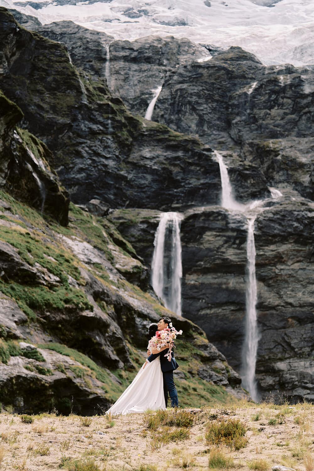 Queenstown New Zealand Heli Wedding Elopement Photographer クイーンズタウン　ニュージーランド　エロープメント 結婚式 | A couple embracing in front of a cascading waterfall on a mountainous terrain, captured by YURI, the Queenstown Wedding Photographer.